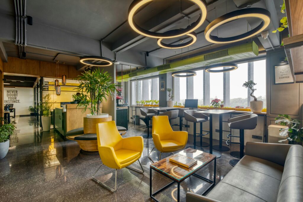 Coworking-lokalets indflydelse på arbejdseffektiviteten 1 Farverigt og moderne kontorrum med gule stole, høje arbejdsborde ved vinduerne, grønne planter og unikke ringformede lamper i loftet.